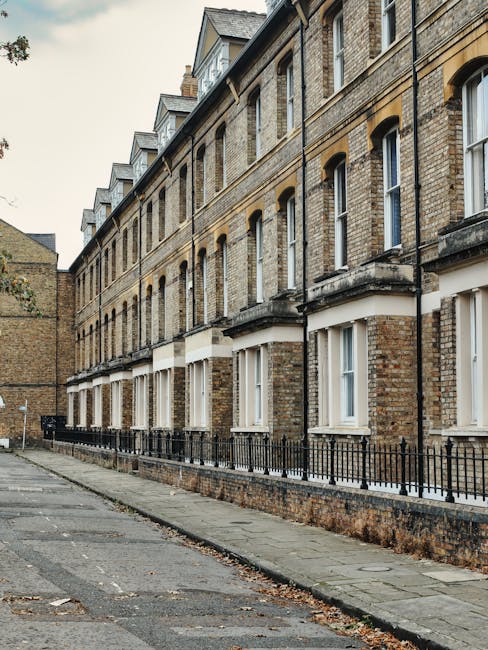 A row of four-storey Victorian terraced houses constructed with yellow and brown brickwork, featuring white-framed sash windows, some with open curtains, and black wrought iron railings along the side