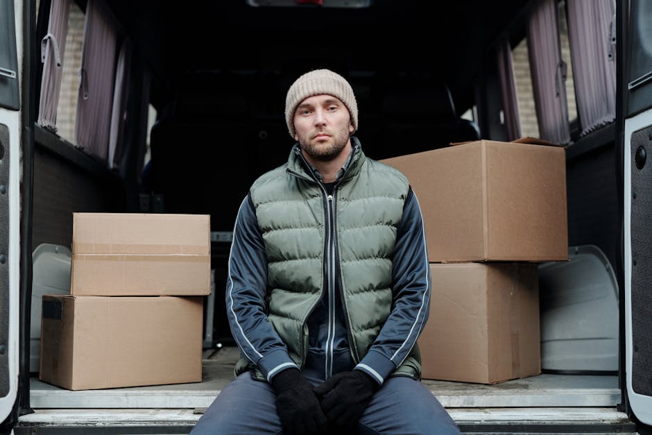 A man wearing a white knit beanie, a green and navy padded jacket, and black gloves stands inside a moving van with the side door open. Behind him are cardboard boxes arranged on the vehicle's floor,