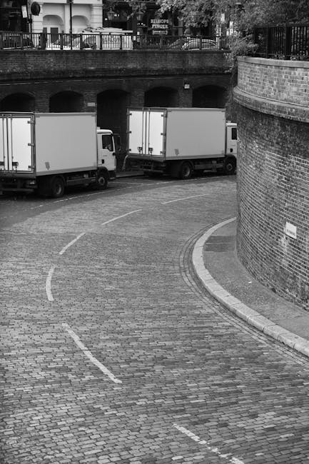 A narrow urban street scene showing parked cars along the curb, with dark-colored vehicles and a white van positioned close to the building on the right side. Several people are visible in the distanc