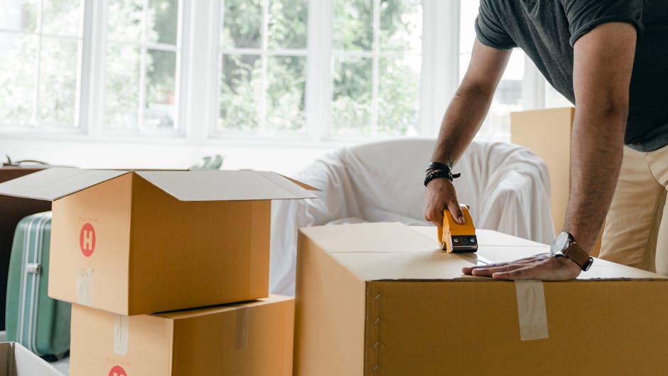 A close-up view of two cardboard boxes used for house removals, placed on a wooden floor inside a property. The larger box in the foreground has white labels with black text reading 'SUMMER CLOTHS' an