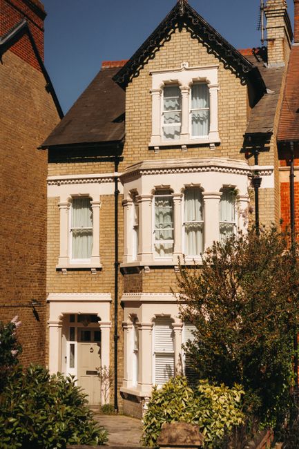 The image shows a row of multi-storey residential buildings on a busy street in Maida Vale, featuring brick facades and large windows. In the foreground, parked cars line the street, with a few pedest