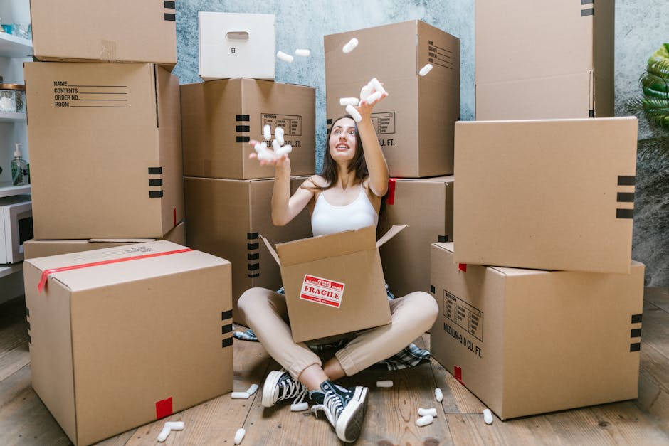 A woman seated on the floor inside a residential property, packing or preparing boxes for house removal, with several cardboard boxes of various sizes stacked near a window and on the carpeted floor i