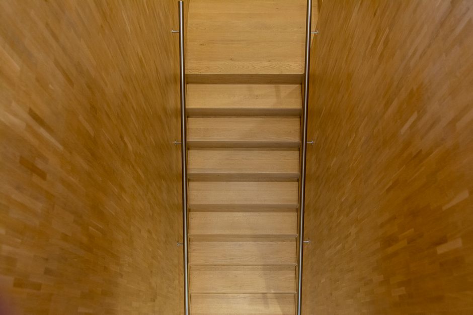 A man wearing a blue and black checked shirt is inside a narrow staircase area of a property, carrying a medium-sized cardboard box. The staircase has black metal railings and beige steps, with a whit